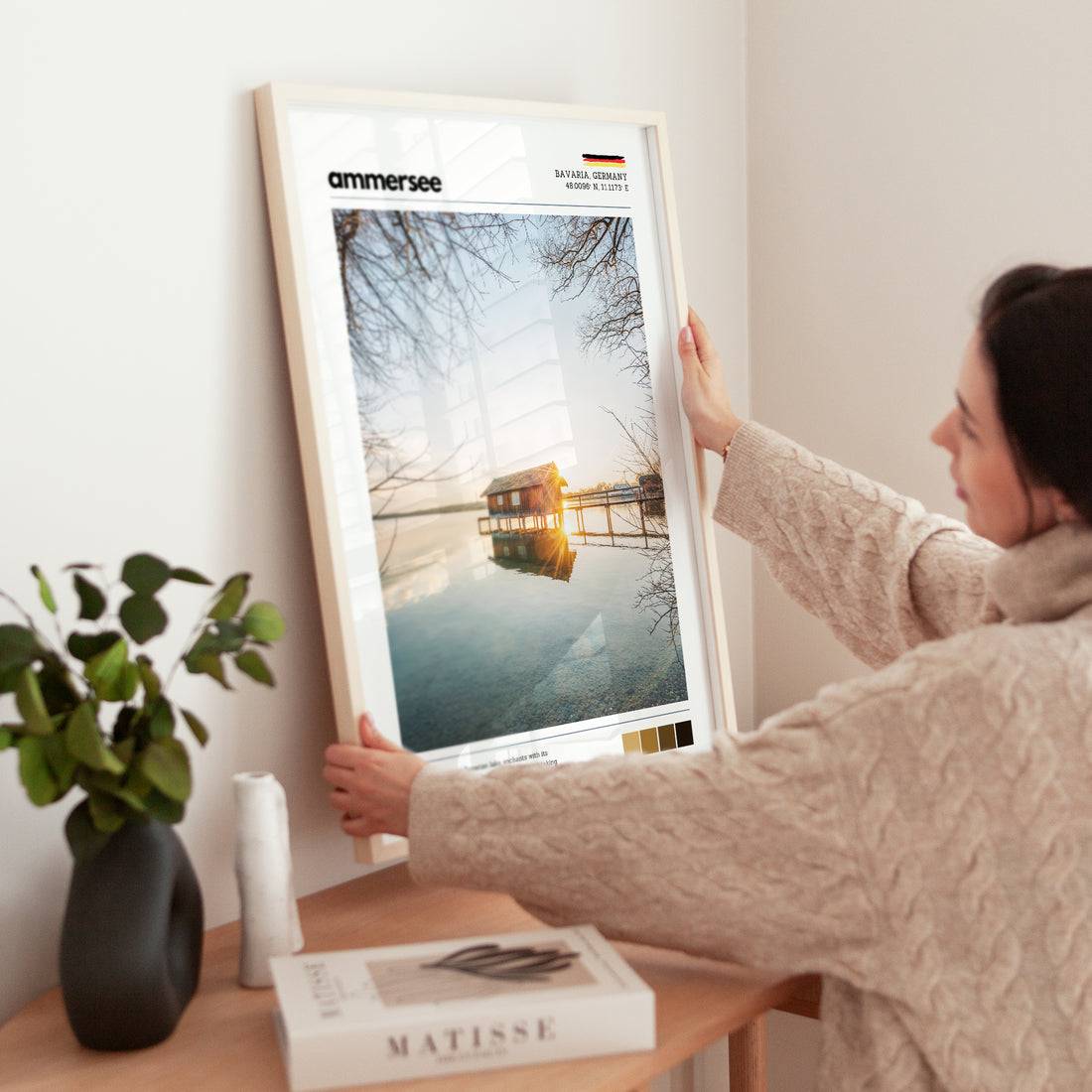 Woman placing large framed Ammersee Bavaria Germany boathouse print in natural wood frame on timber sideboard beside plant and books