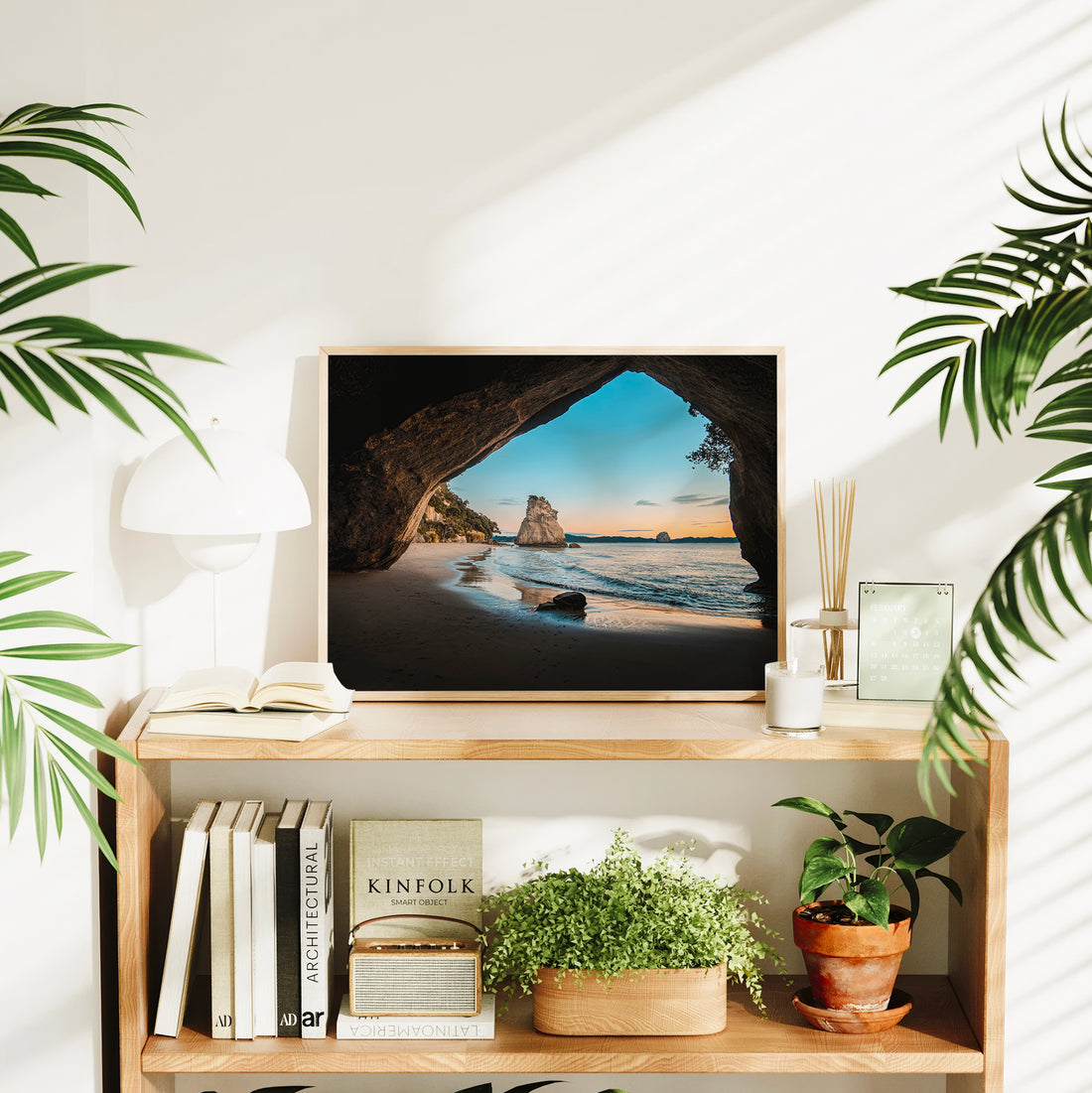 Natural wood framed Cathedral Cove sunrise landscape print displayed on wooden bookshelf in bright interior styled with plants and books