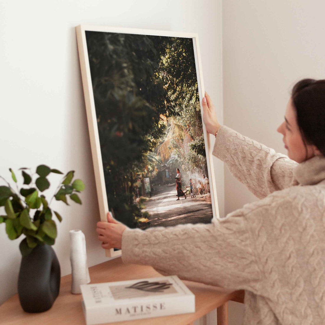 Bali village street photography print with golden jungle canopy light in natural wood frame leaning against peach terracotta wall