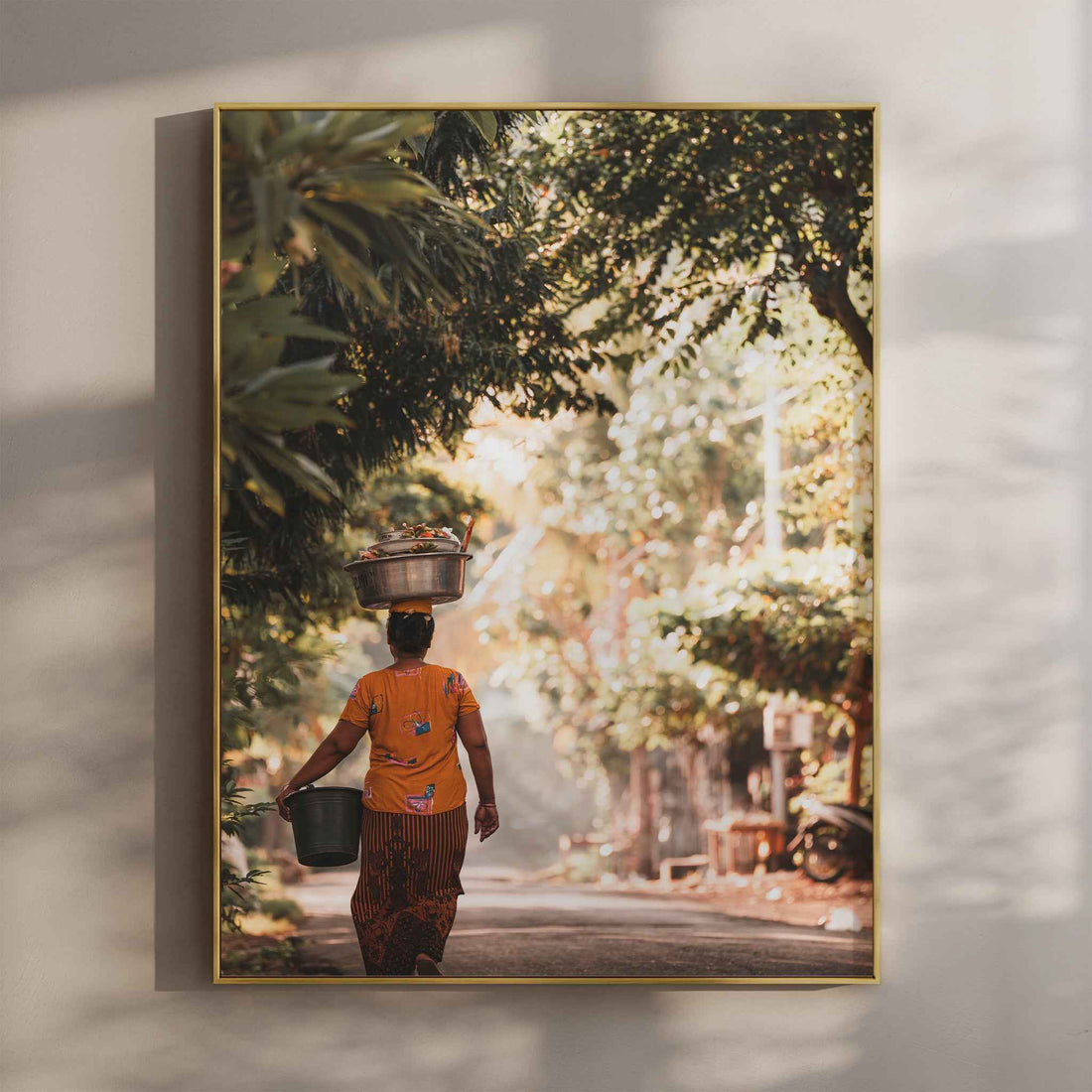 Balinese woman carrying offerings on head through jungle village road wall art print in gold floating frame mounted on beige wall with natural shadows