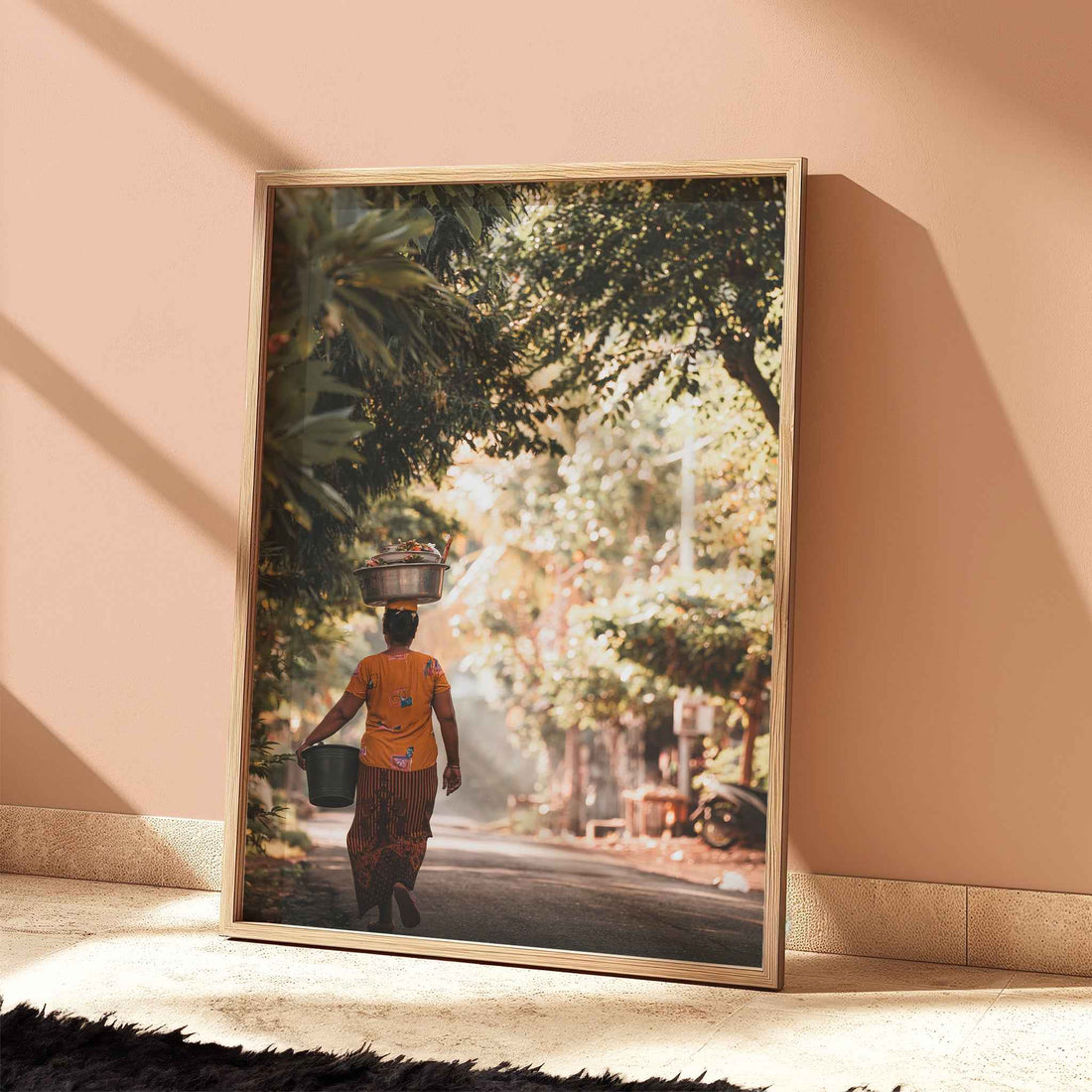 Balinese woman carrying offerings through golden jungle road Bali print in natural wood frame leaning against peach terracotta wall
