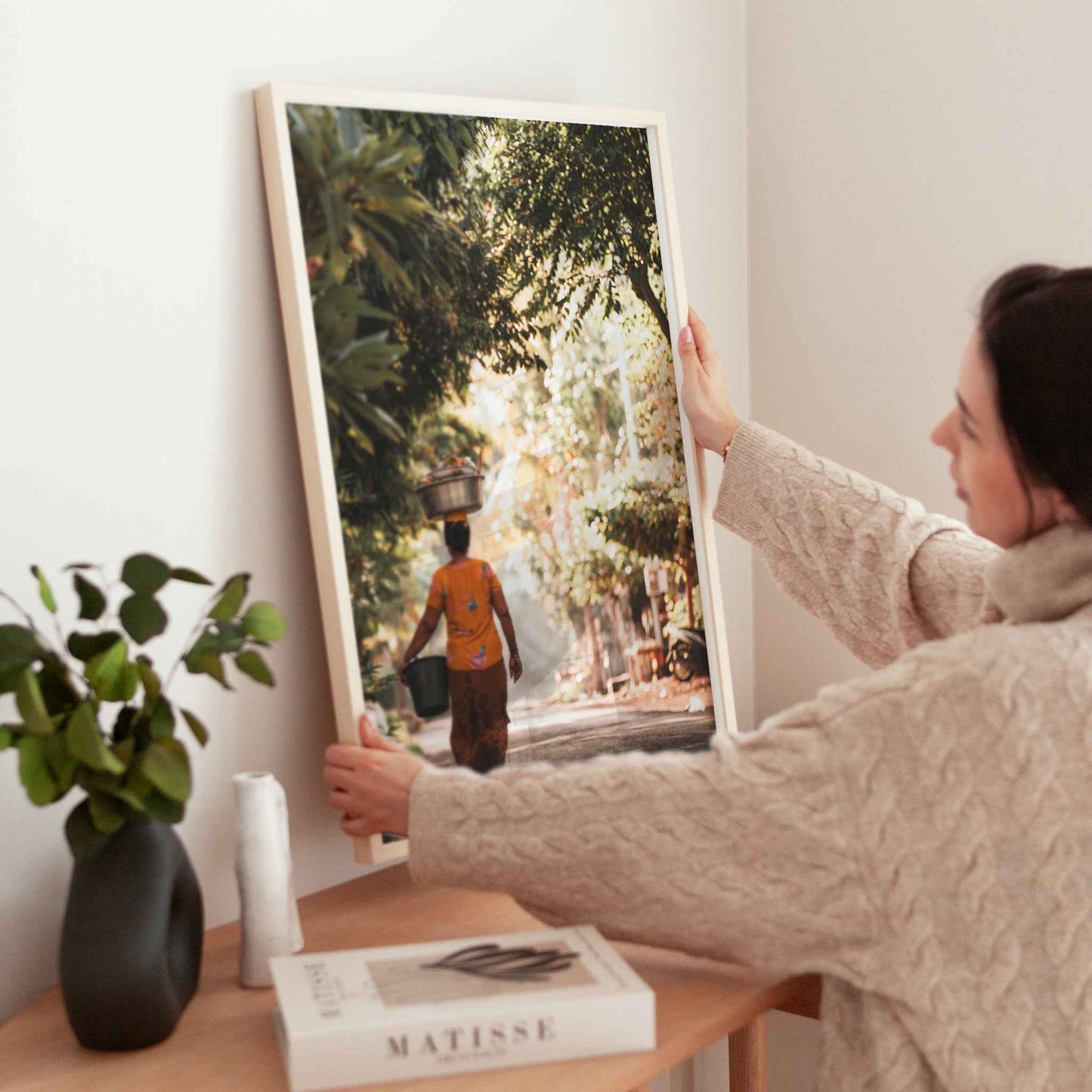 Woman holding large Balinese woman carrying offerings framed print in natural wood frame above wooden side table with Matisse book and plant