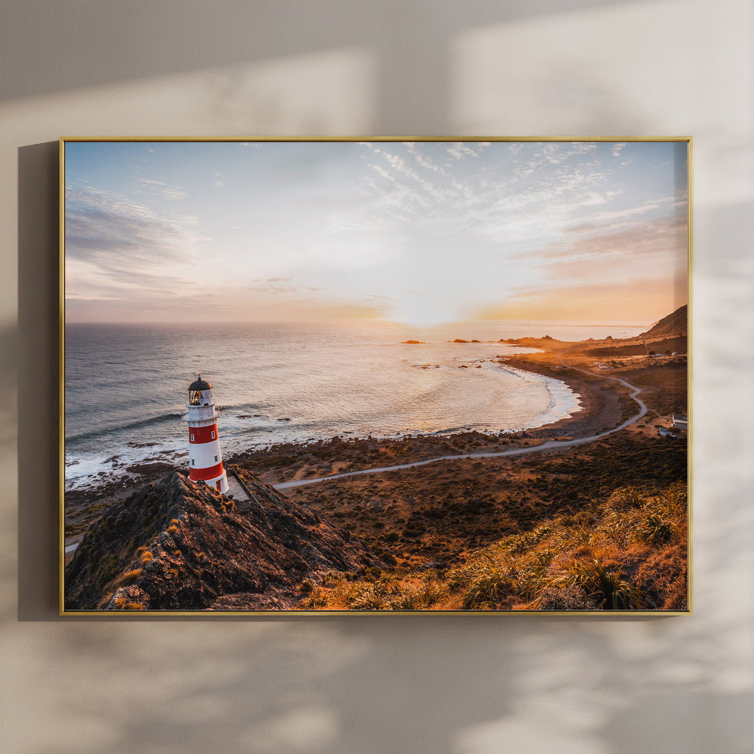 Framed Cape Palliser lighthouse sunset landscape photography print in gold float frame mounted on warm beige wall with window light shadows