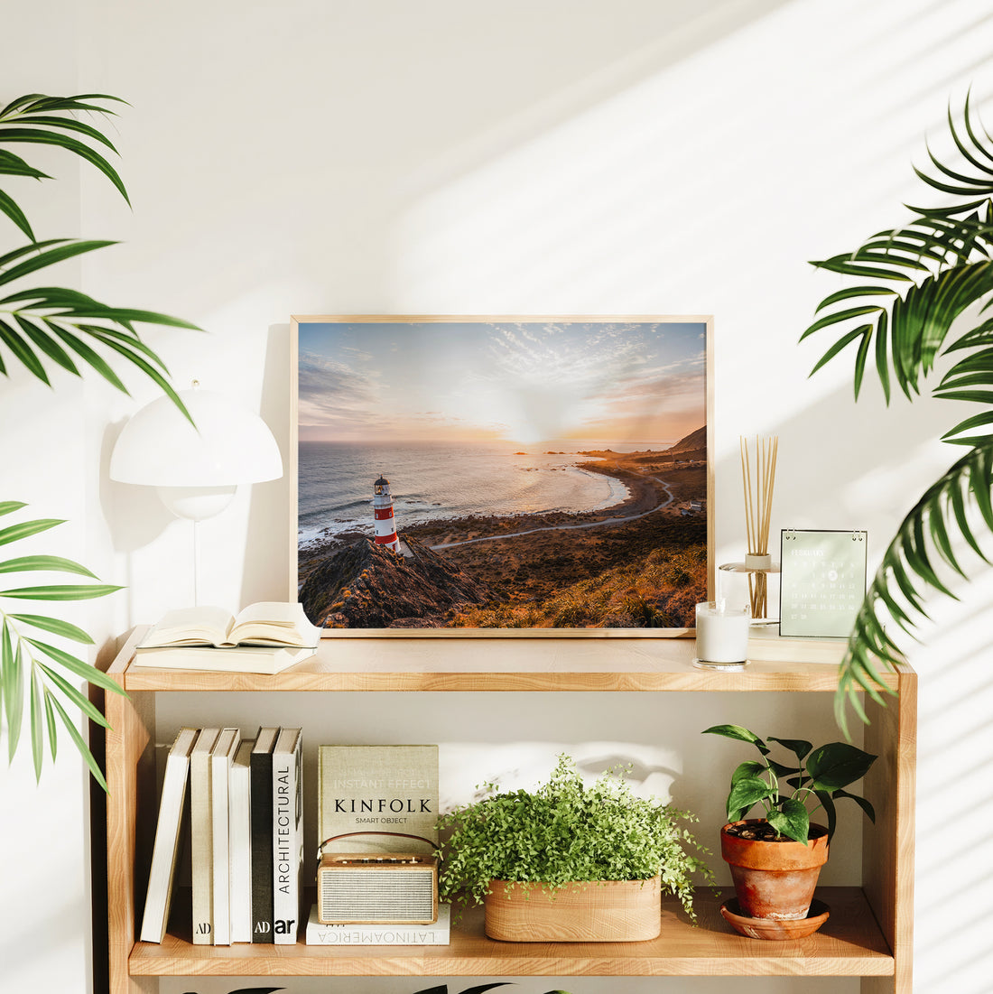Natural wood framed Cape Palliser lighthouse landscape print displayed on wooden bookshelf in bright interior styled with plants and books