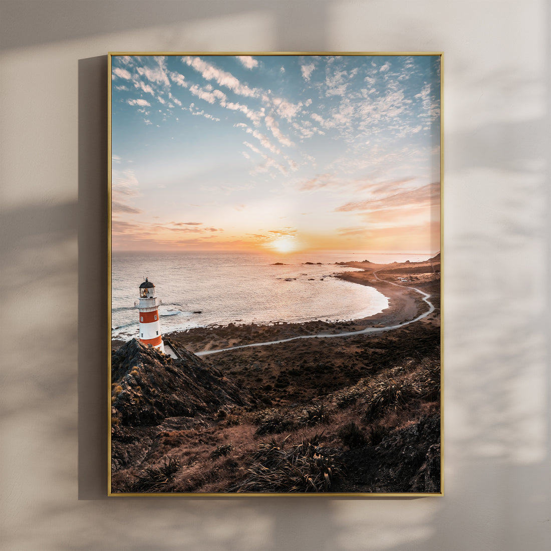 Framed Cape Palliser lighthouse sunset photography print in gold float frame mounted on warm beige wall with window light shadows