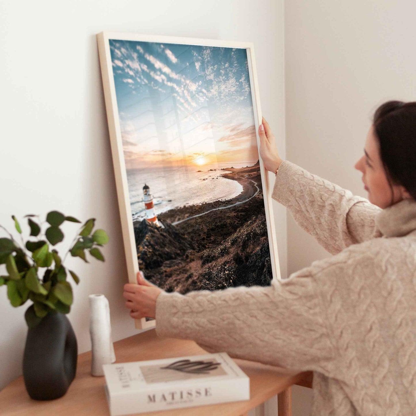 Woman holding large framed Cape Palliser lighthouse sunset photography print in natural wood frame near wooden table with plant and books