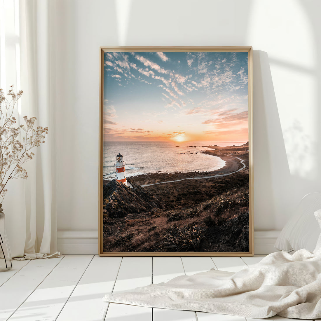 Natural wood framed Cape Palliser lighthouse sunset print leaning against white wall on white floorboards in minimal Scandinavian interior