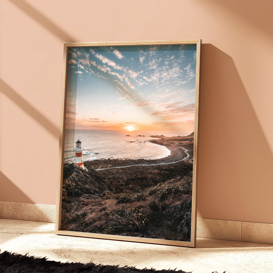 Framed Cape Palliser lighthouse sunset print in light wood frame leaning against terracotta wall with dramatic shadows