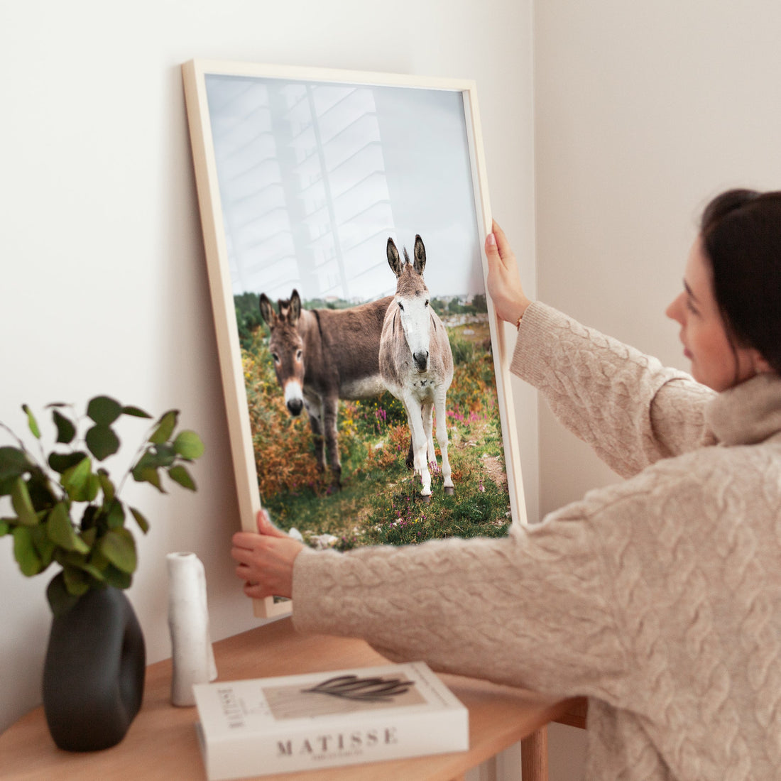 Woman adjusting a framed donkey art print on a wooden table.