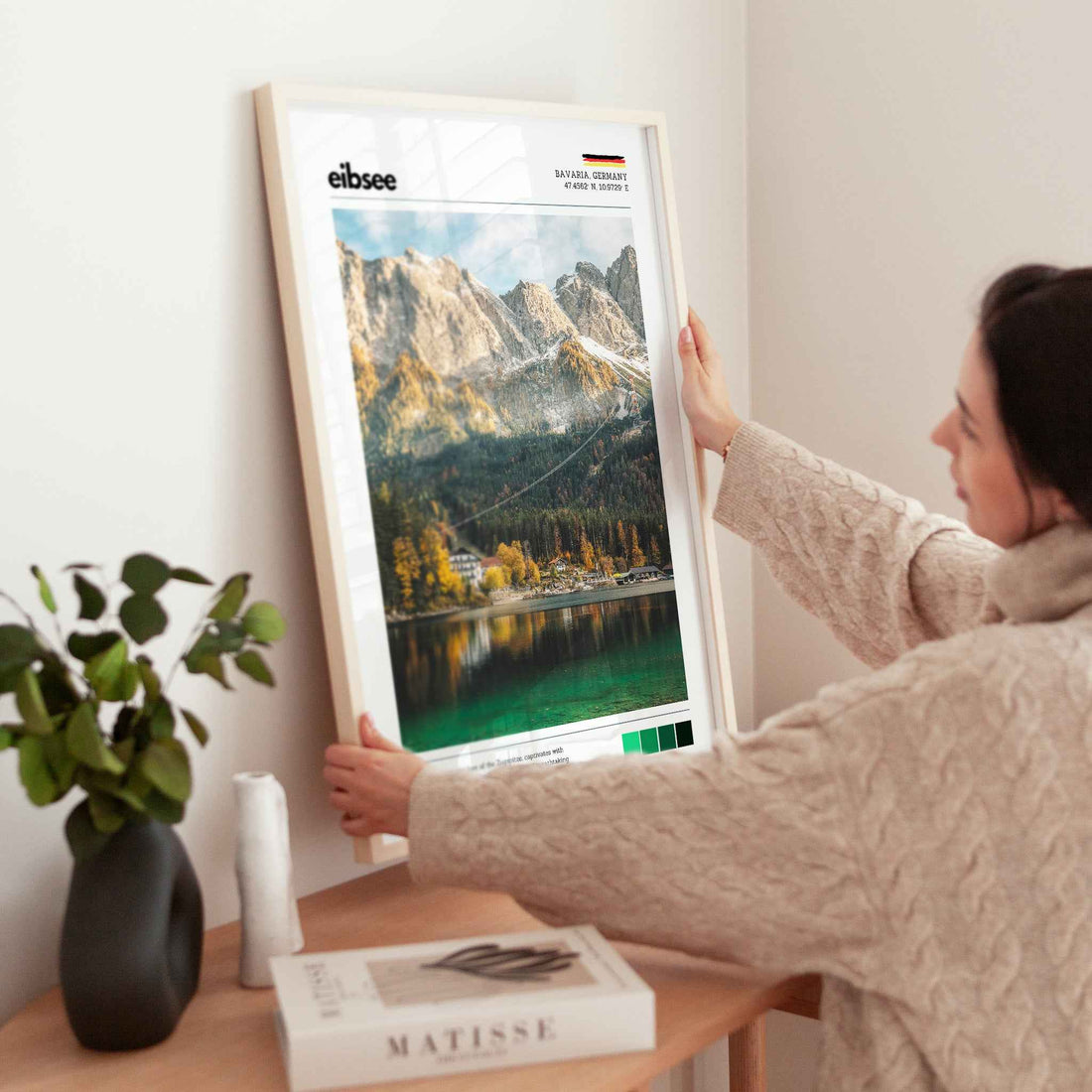 Woman placing large framed Eibsee autumn Zugspitze Bavaria Germany print in natural wood frame on timber sideboard beside plant and books