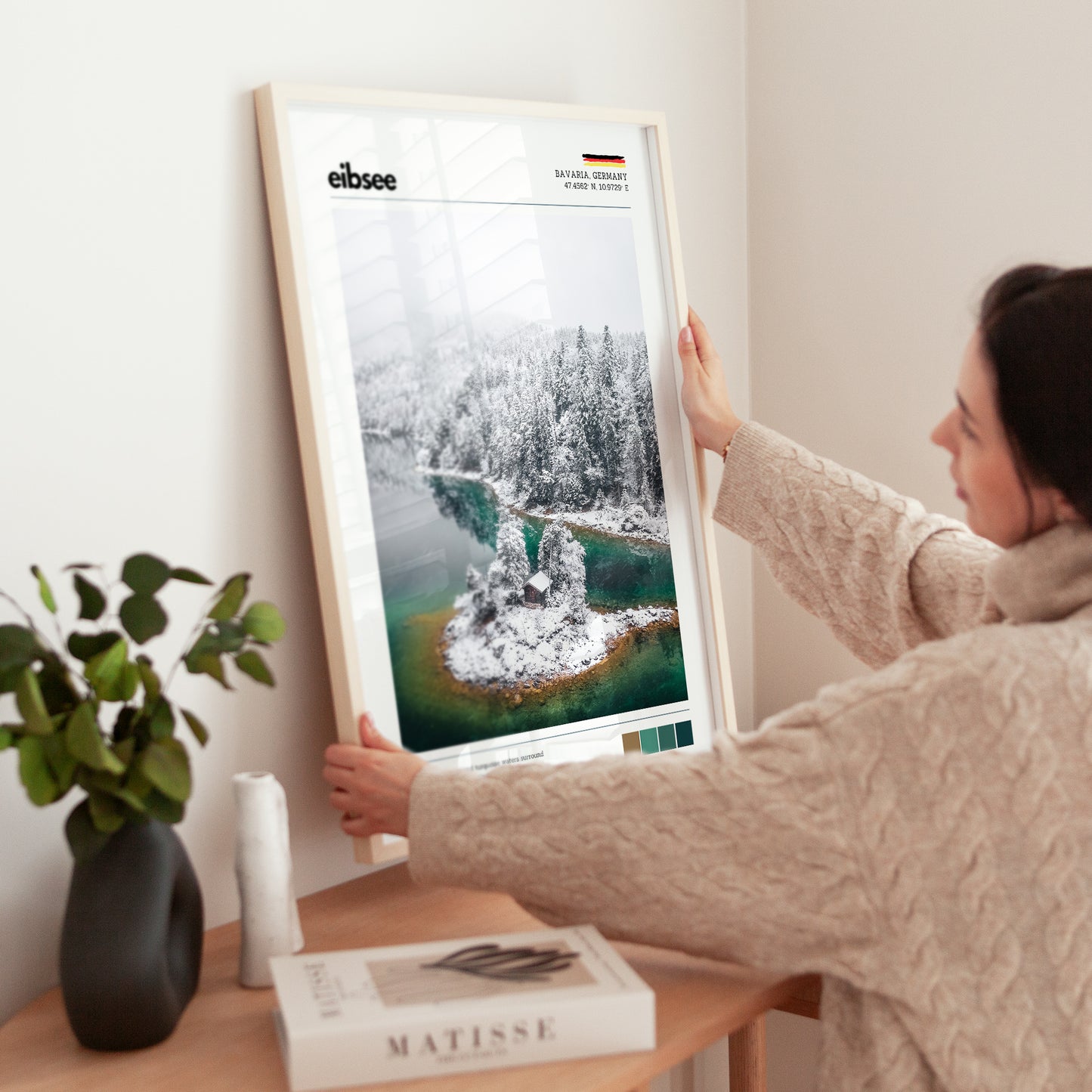Woman placing large framed Eibsee winter island cabin Bavaria Germany print in natural wood frame on timber sideboard beside plant and books