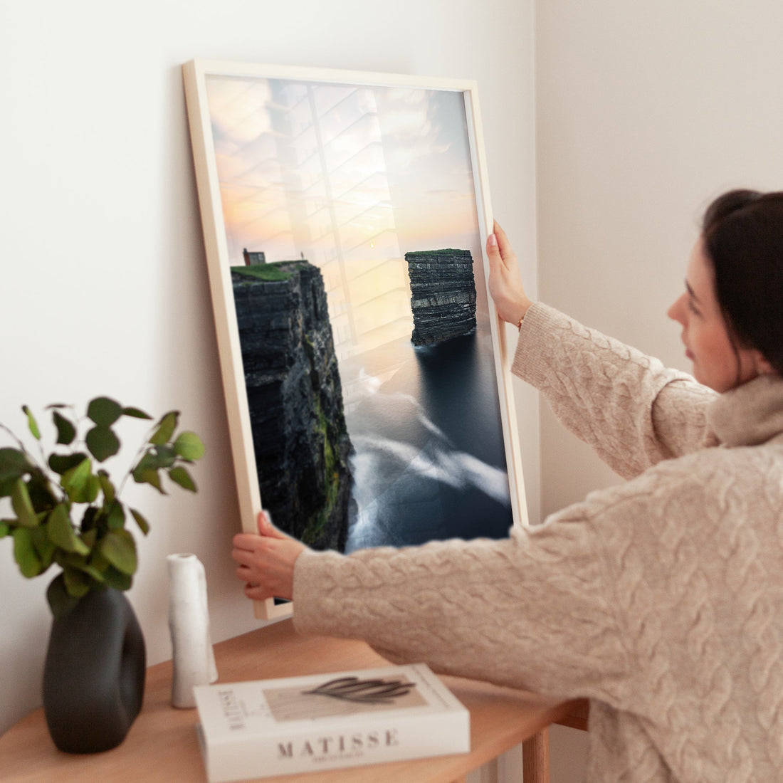 Woman positioning framed Downpatrick Head wall art print in a minimal, modern room.