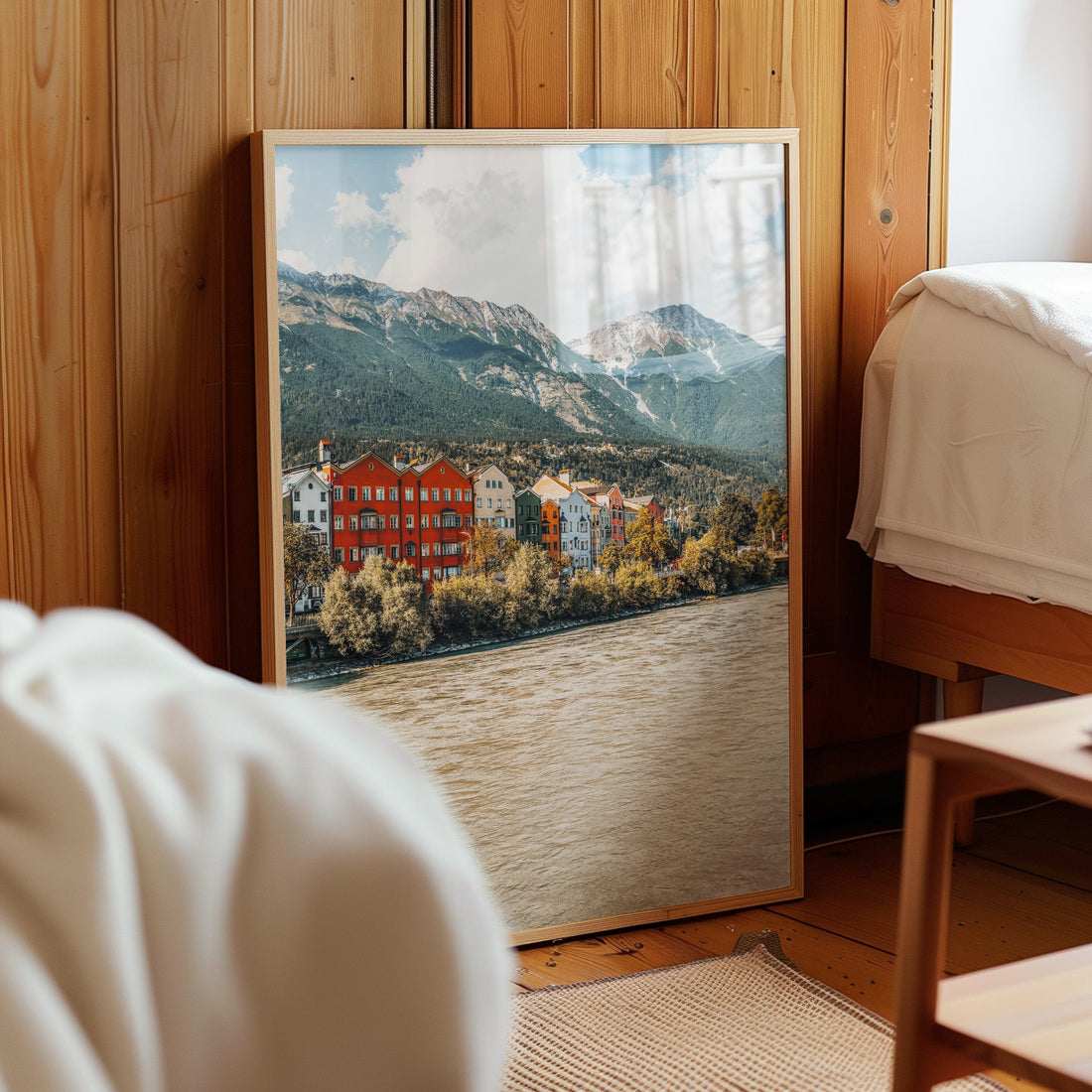 Framed Innsbruck mountain and river print leaning against wood wall in warm bedroom.