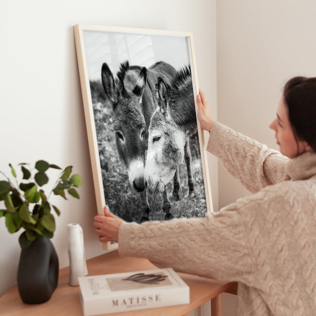 Woman holding framed black and white donkey art on wooden dresser in a cosy home.