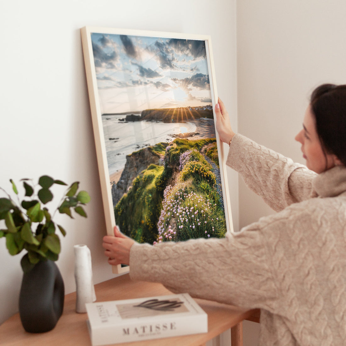 Woman placing framed coastal print of Annestown with sunset glow and vibrant cliffside flowers.