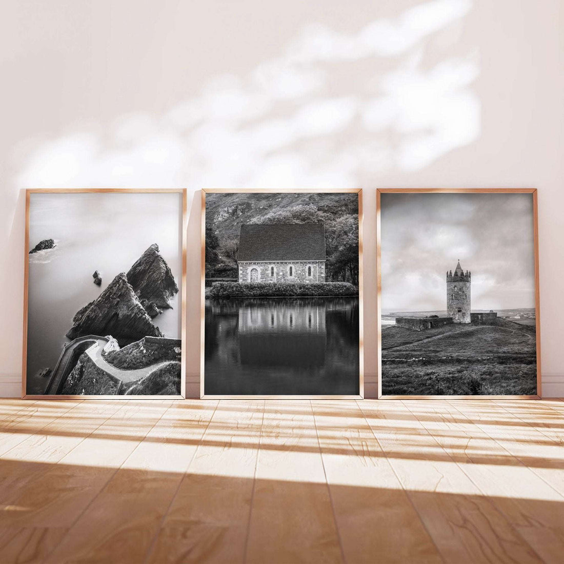 Three black and white Irish landscape prints in light wooden frames, leaning against a wall on a wooden floor, featuring a coastal bothy, Gougane Barra church, and a stone tower.