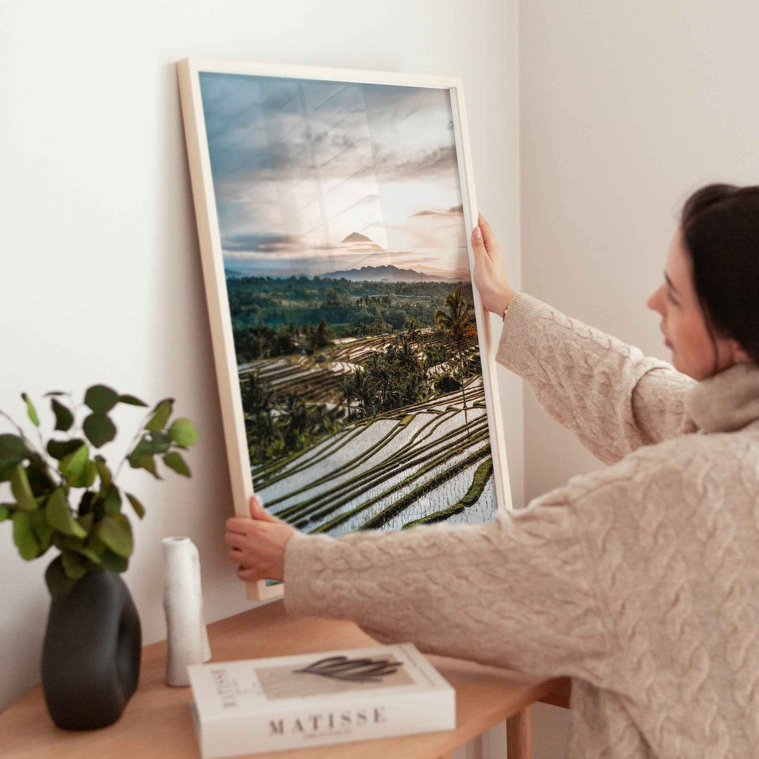 Woman holding large Jatiluwih rice terraces Bali framed print in natural wood frame above wooden side table with Matisse book and plant