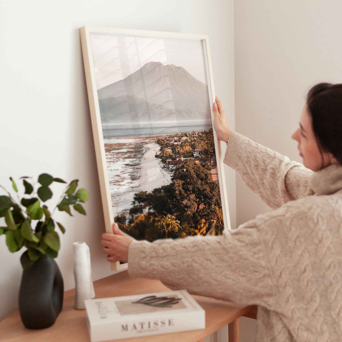 Woman holding large Jungutbatu Beach Nusa Lembongan framed print in natural wood frame above wooden side table with Matisse book and plant