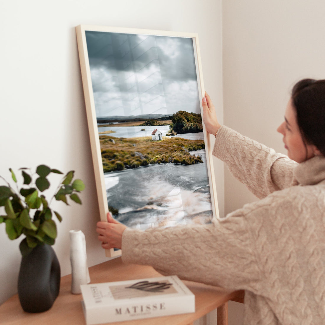 Woman positioning a framed print of a white Connemara hut by a rushing river