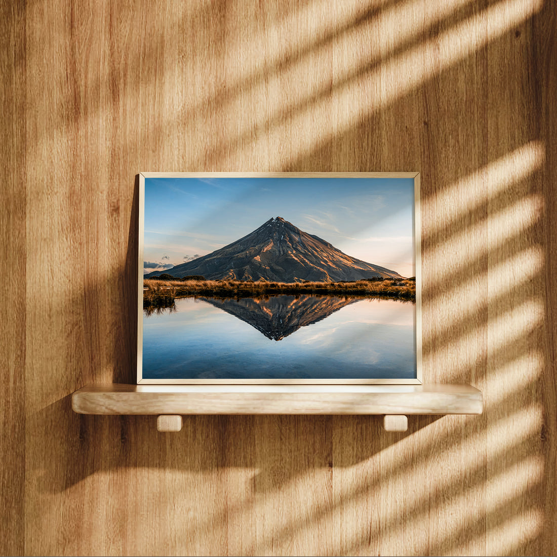 Framed Mount Taranaki volcano reflection print in natural wood frame leaning on shelf against warm oak panelled wall