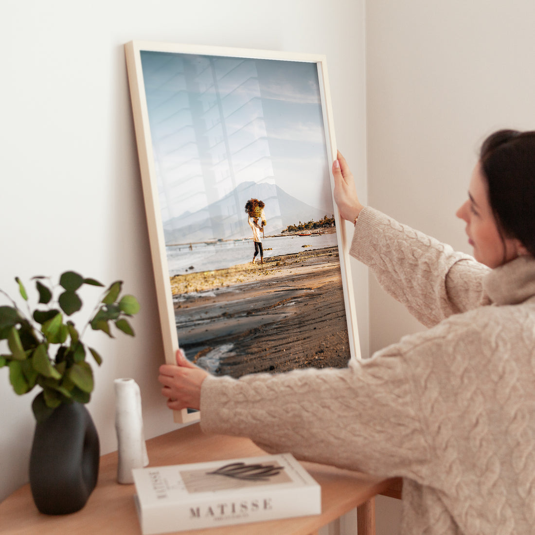 Woman holding large Nusa Lembongan seaweed farmer framed print in natural wood frame above wooden side table with Matisse book and plant