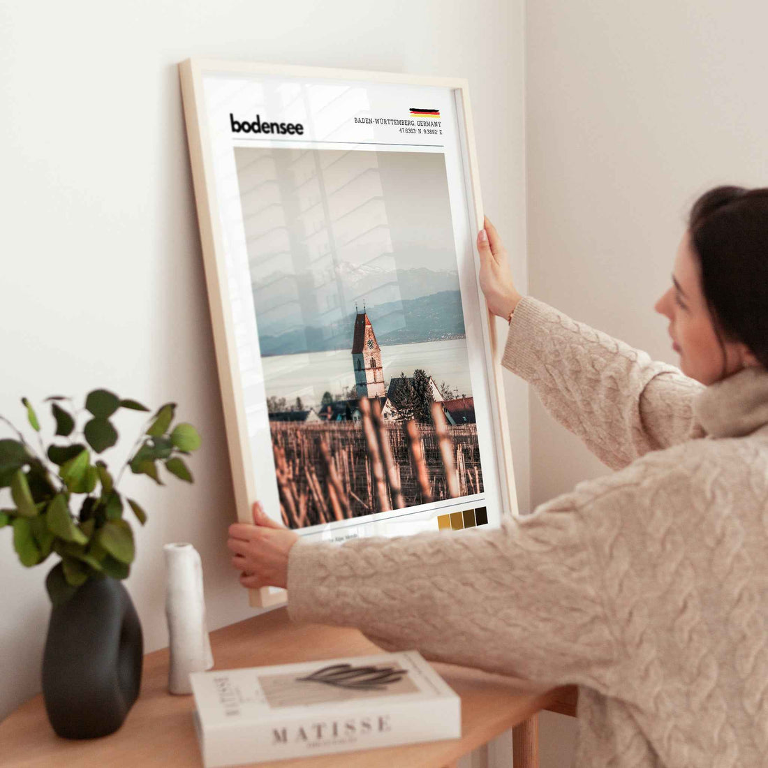 Woman placing large framed Bodensee Lake Constance Germany church vineyard print in natural wood frame on timber sideboard beside plant and books