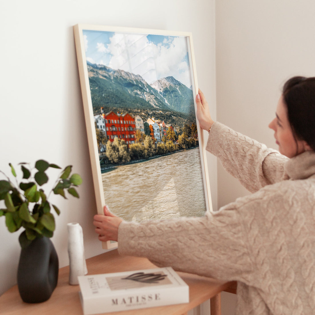 Woman styling home with Innsbruck landscape print of colourful houses by river and mountains.