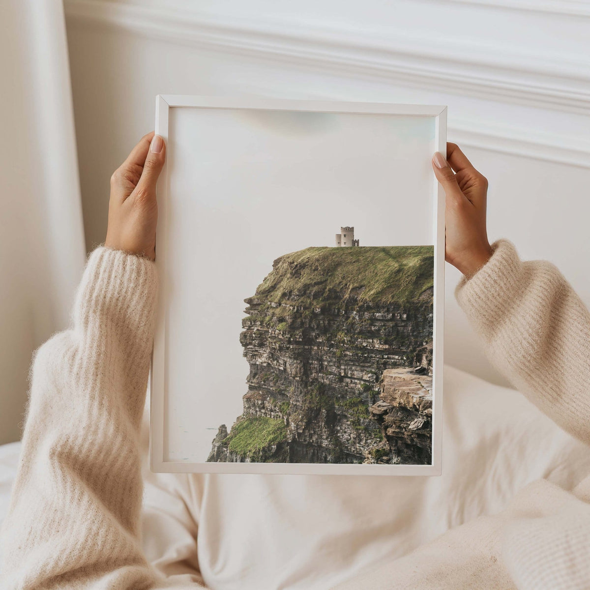 Hands holding a white-framed Cliffs of Moher print showing rocky coastal cliffs, displayed in a bright white interior