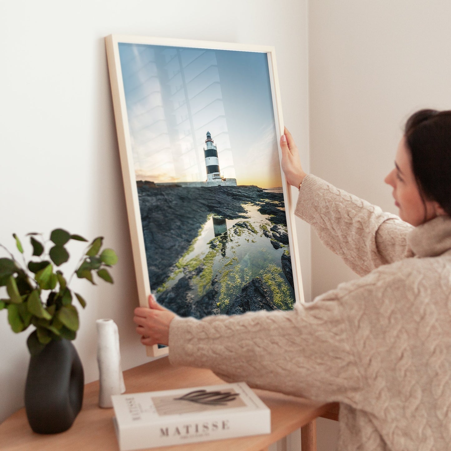 Woman placing framed lighthouse print on table, warm and minimal interior styling