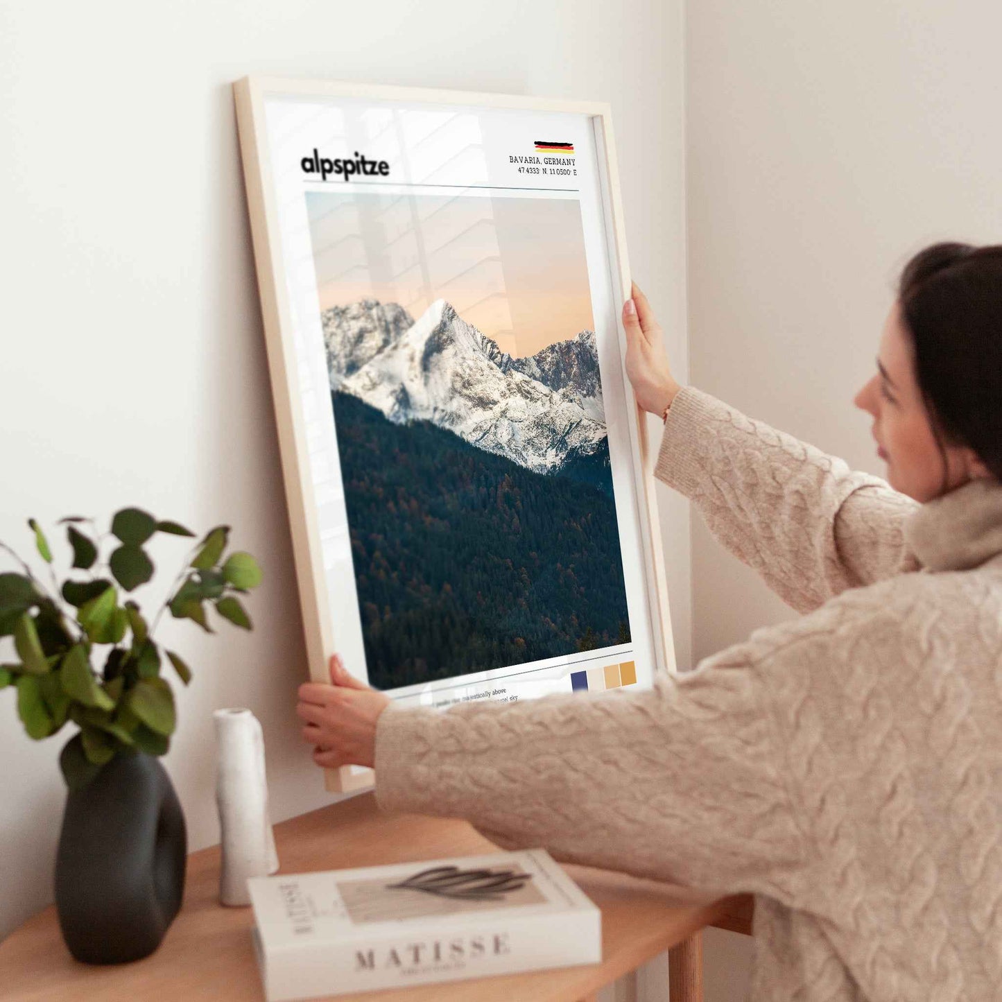 Woman placing large framed Alpspitze Bavaria Germany print in natural wood frame on timber sideboard beside plant and books
