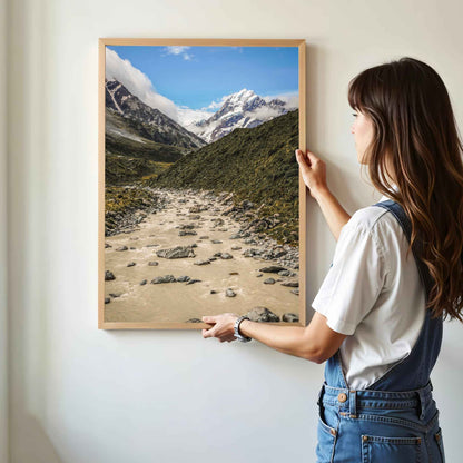Woman hanging a natural wood framed Aoraki Mount Cook Hooker Valley New Zealand print on a white wall