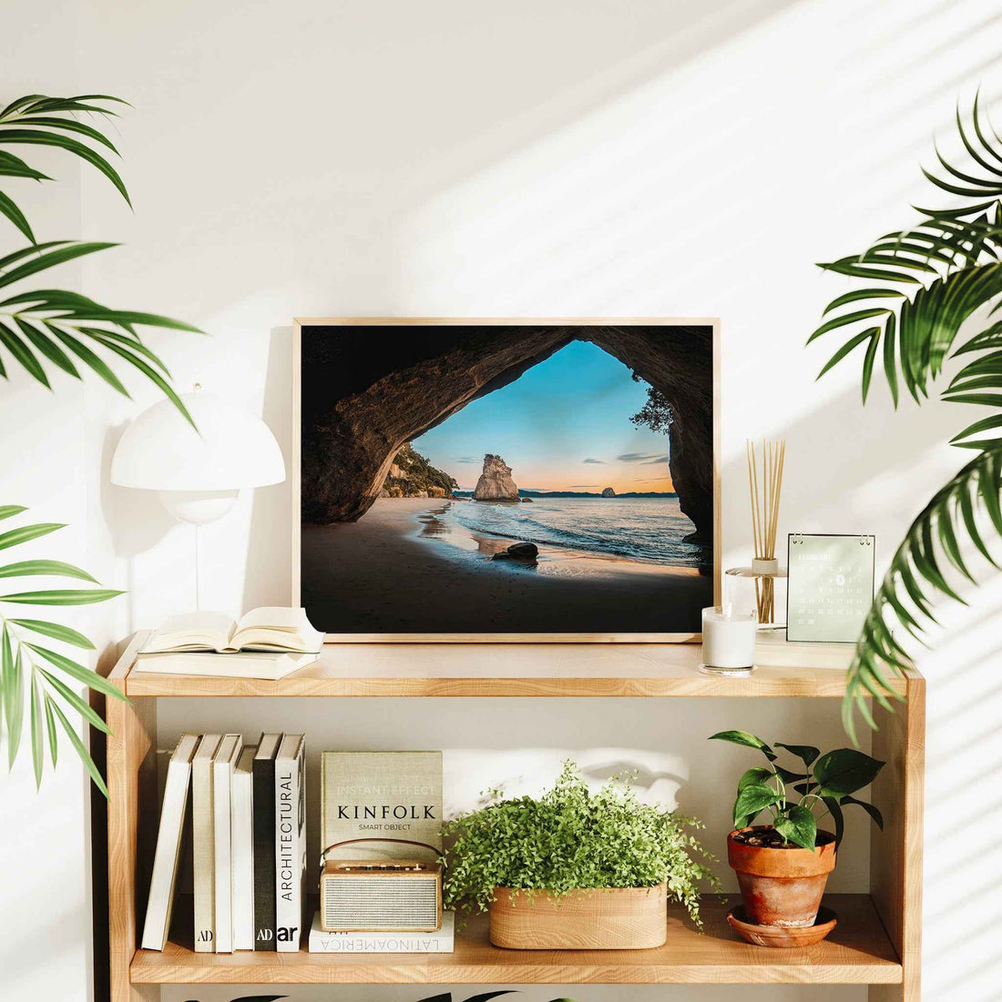 Natural wood framed Cathedral Cove sunrise landscape print displayed on wooden bookshelf in bright interior styled with plants and books