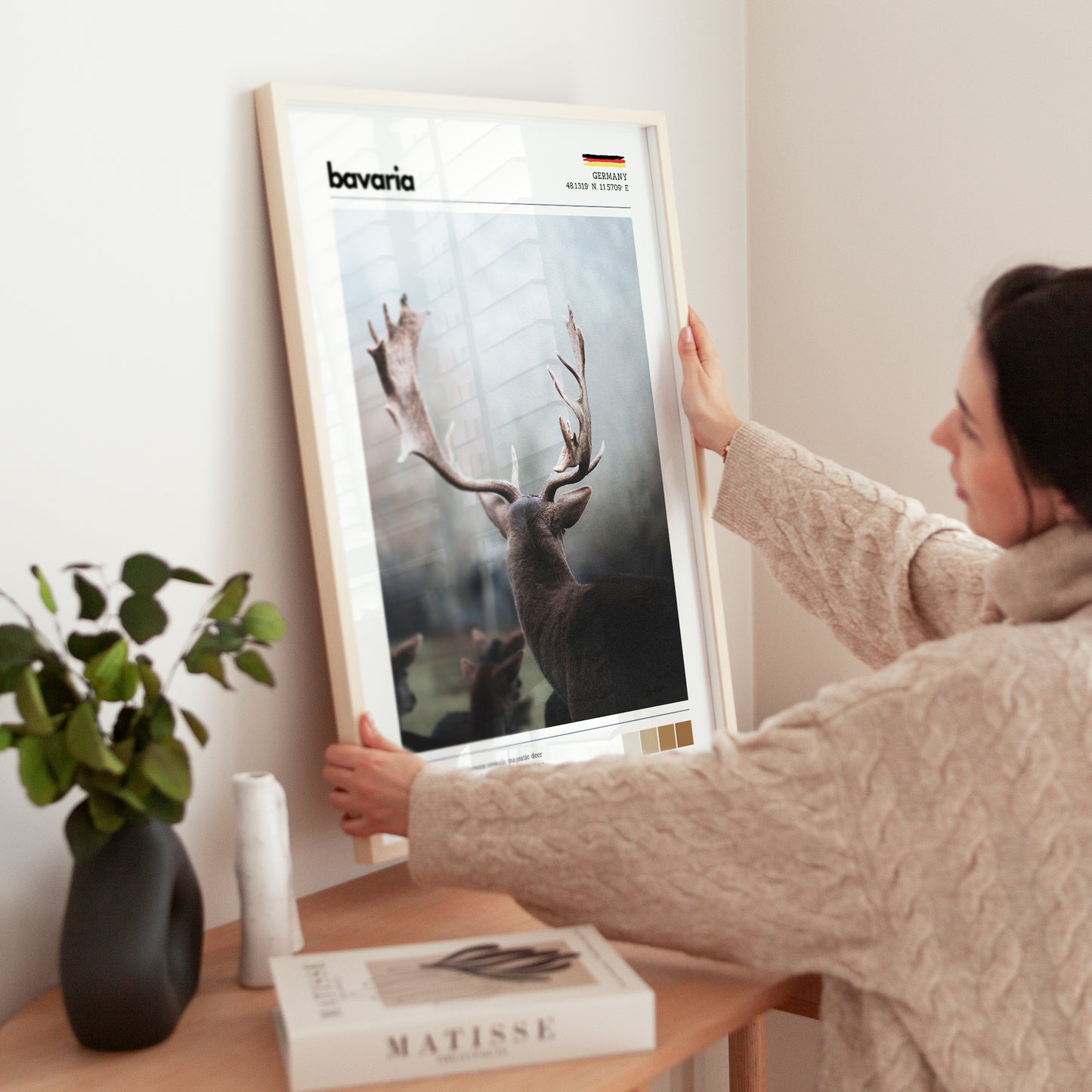 Woman placing large framed Bavaria red deer stag Germany print in natural wood frame on timber sideboard beside plant and books