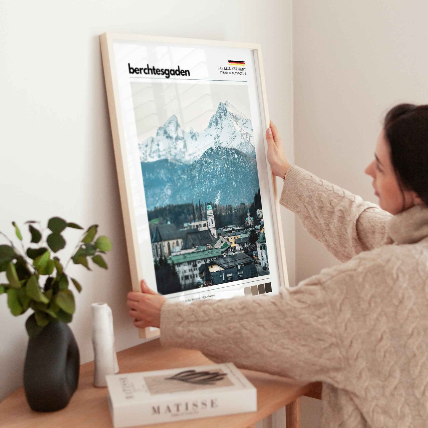 Woman placing large framed Berchtesgaden Bavaria Germany town and Watzmann print in natural wood frame on timber sideboard beside plant and books
