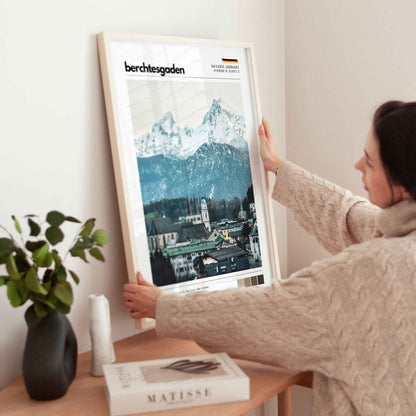 Woman placing large framed Berchtesgaden Bavaria Germany town and Watzmann print in natural wood frame on timber sideboard beside plant and books