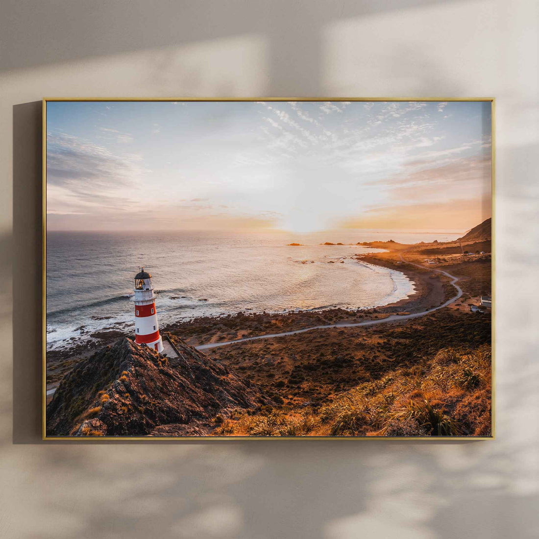 Framed Cape Palliser lighthouse sunset landscape photography print in gold float frame mounted on warm beige wall with window light shadows
