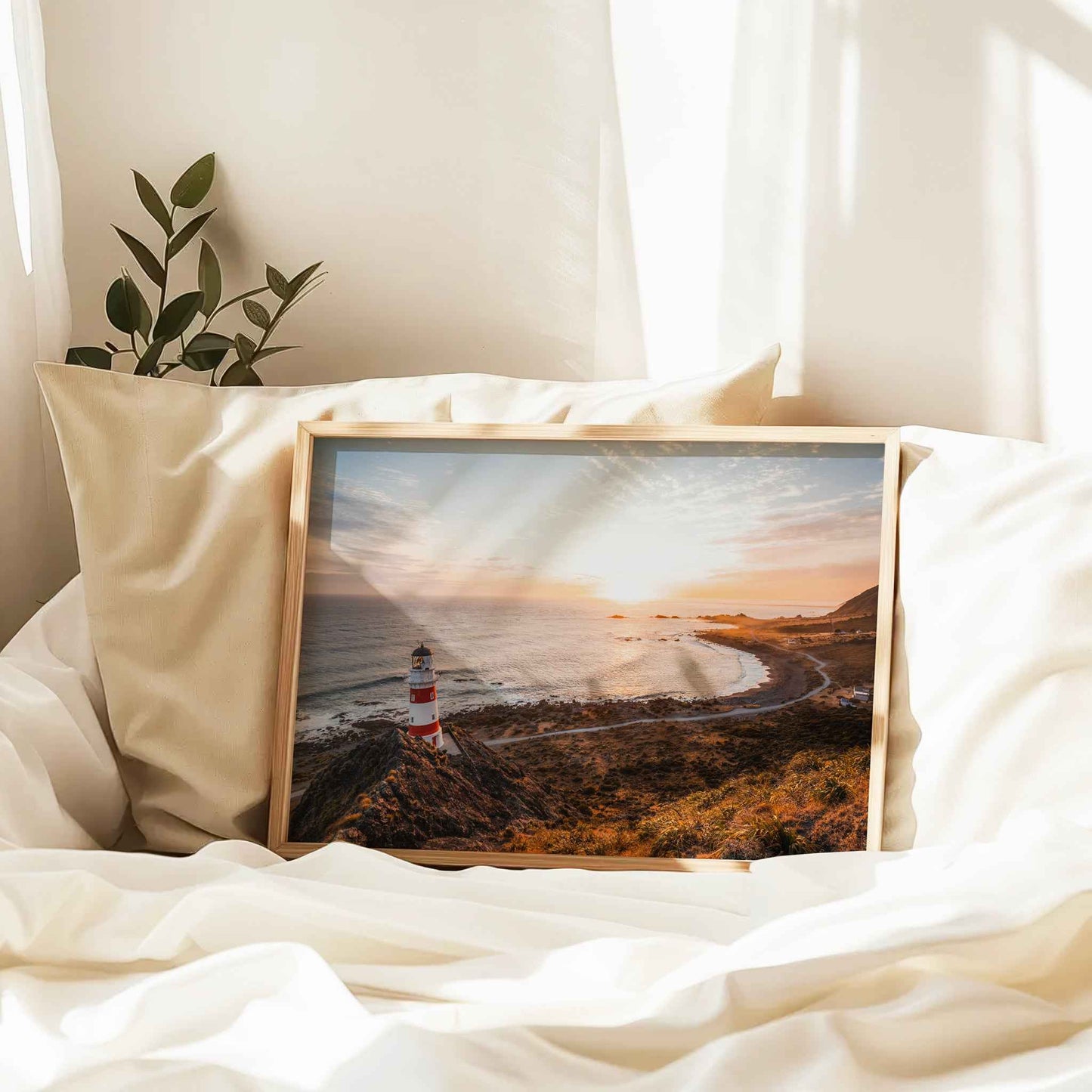 Framed Cape Palliser lighthouse landscape print in natural wood frame resting against linen pillows on white bed with plant and curtains