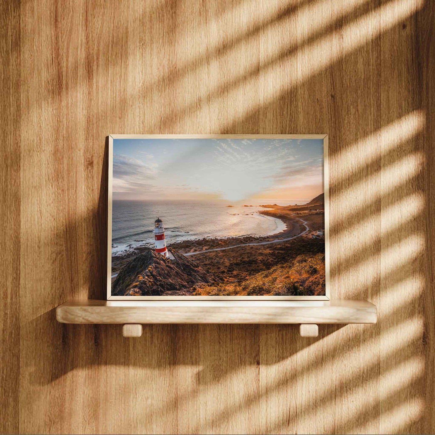 Framed Cape Palliser lighthouse landscape print in natural wood frame displayed on wooden shelf against warm oak panelled wall with dappled light