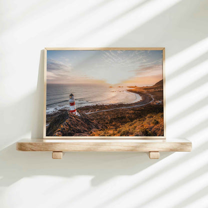 Framed Cape Palliser lighthouse landscape photography print in natural wood frame on minimalist wooden shelf against white wall with dappled shadows