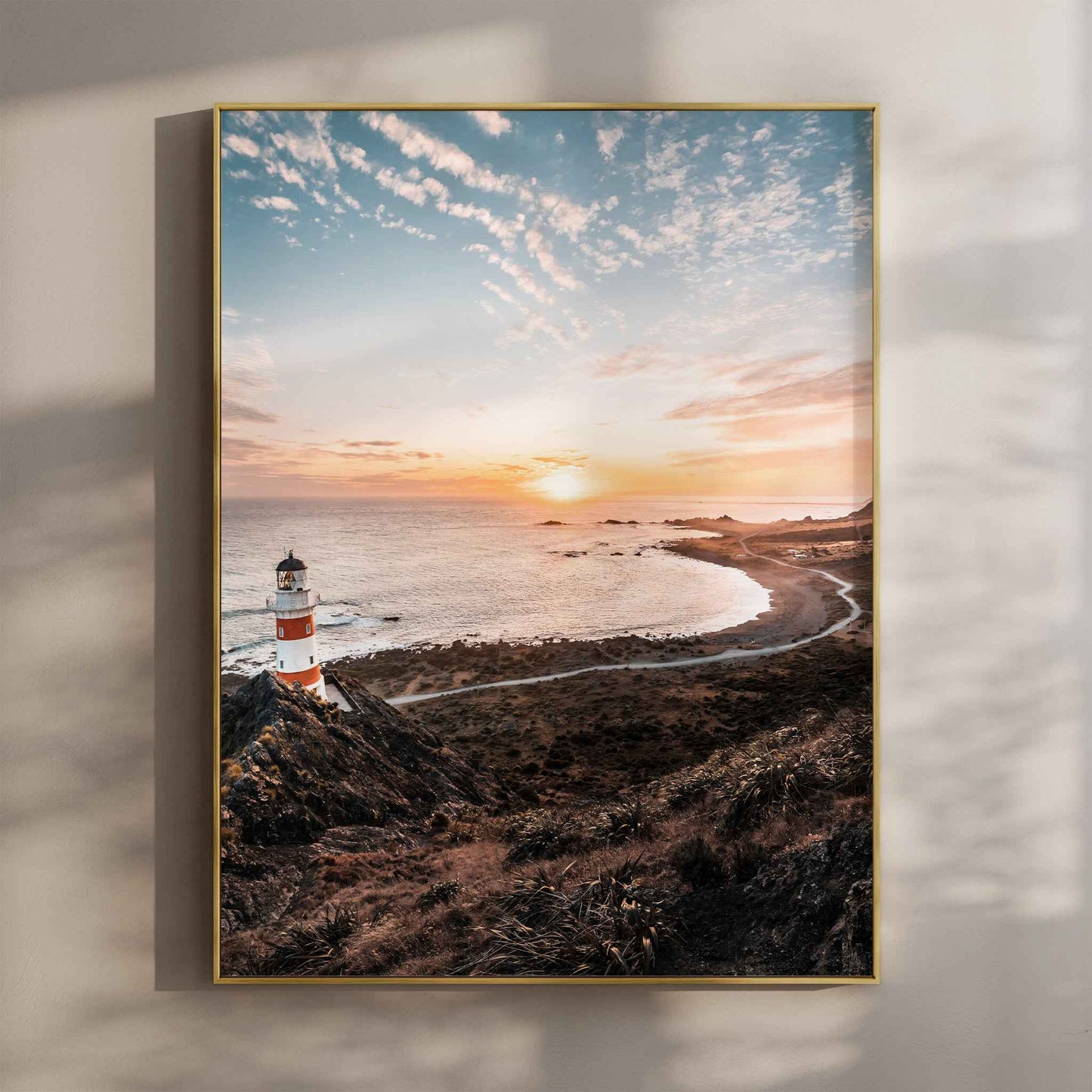 Framed Cape Palliser lighthouse sunset photography print in gold float frame mounted on warm beige wall with window light shadows