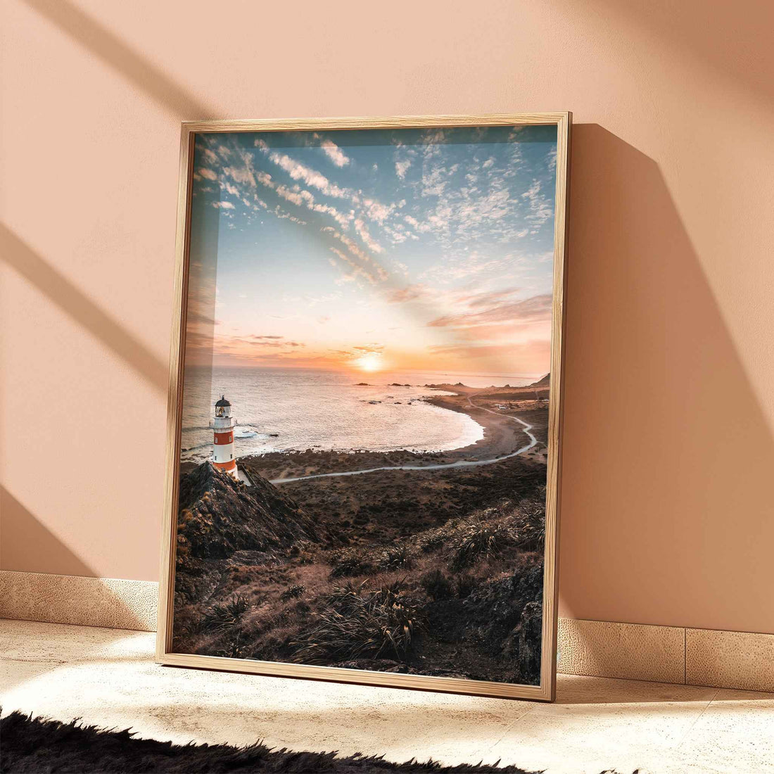 Framed Cape Palliser lighthouse sunset print in light wood frame leaning against terracotta wall with dramatic shadows