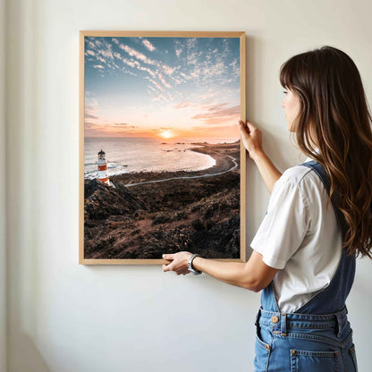 Woman hanging framed Cape Palliser lighthouse sunset photography print in natural oak frame on white wall