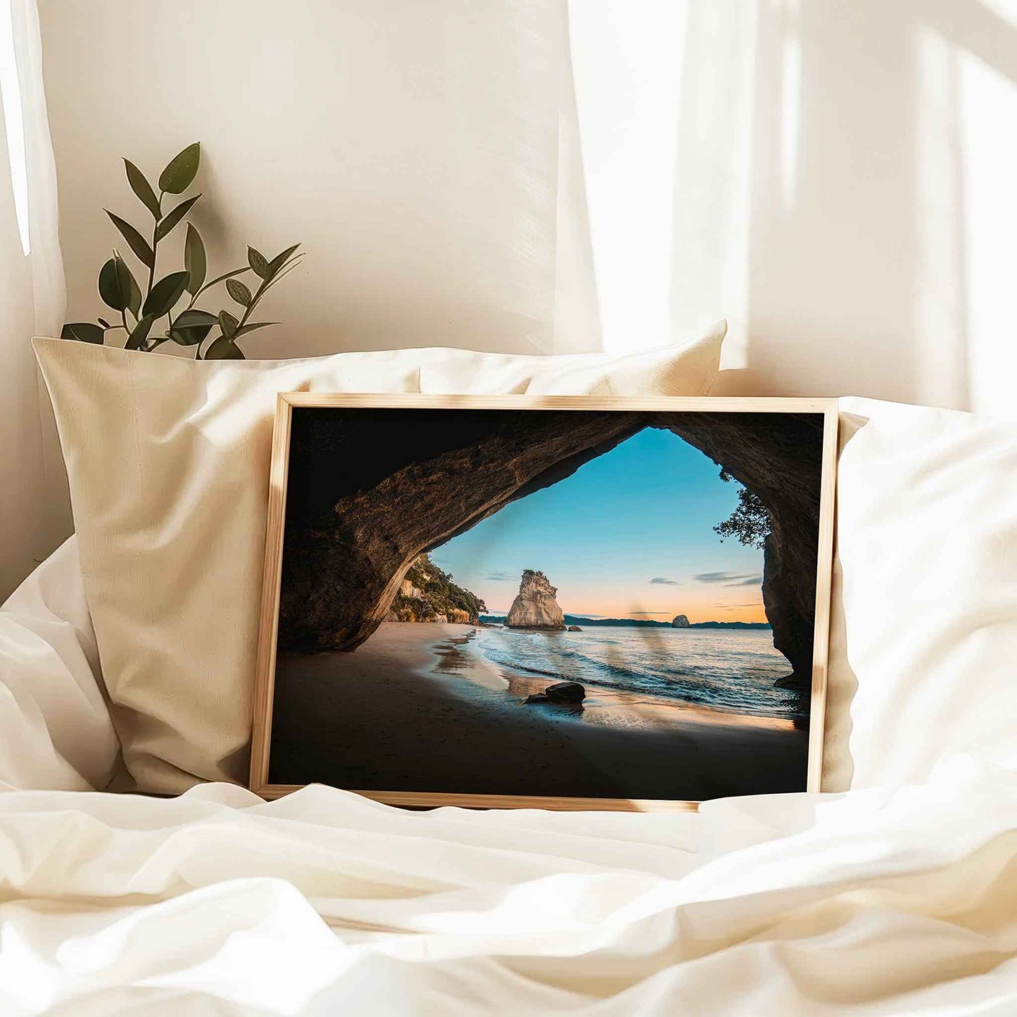 Framed Cathedral Cove sunrise landscape print in natural wood frame resting against linen pillows on white bed with plant and curtains