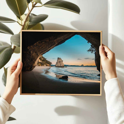 Hands holding framed Cathedral Cove sunrise landscape photography print in natural oak frame against white wall with large leaf plant