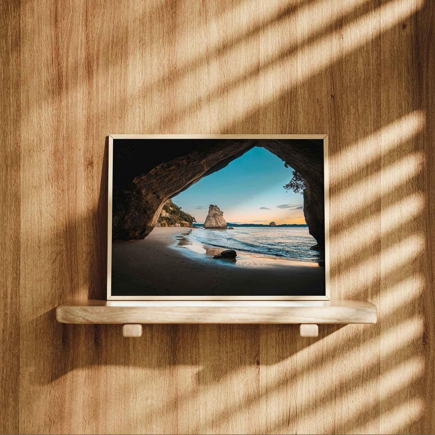Framed Cathedral Cove sunrise landscape print in natural wood frame displayed on wooden shelf against warm oak panelled wall with dappled light
