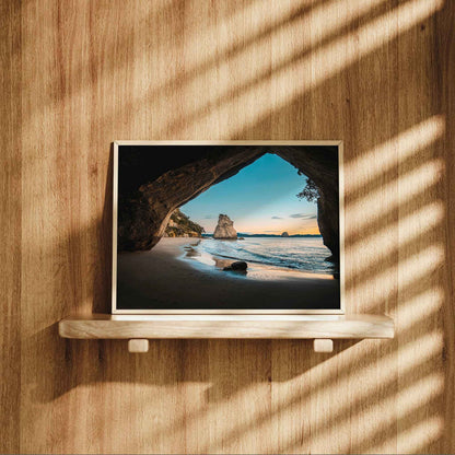 Framed Cathedral Cove sunrise landscape print in natural wood frame displayed on wooden shelf against warm oak panelled wall with dappled light
