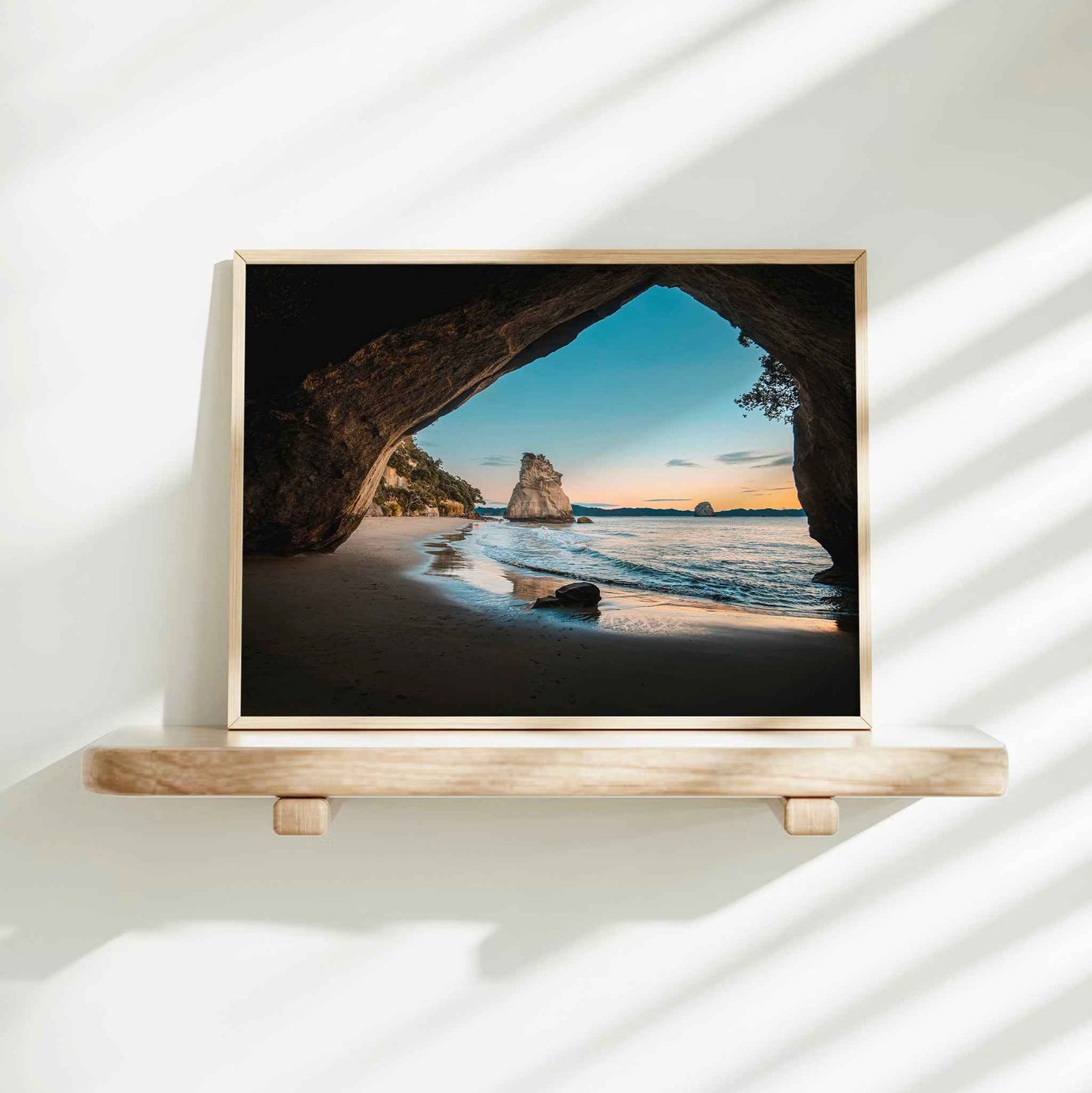 Framed Cathedral Cove sunrise landscape photography print in natural wood frame on minimalist wooden shelf against white wall with dappled shadows