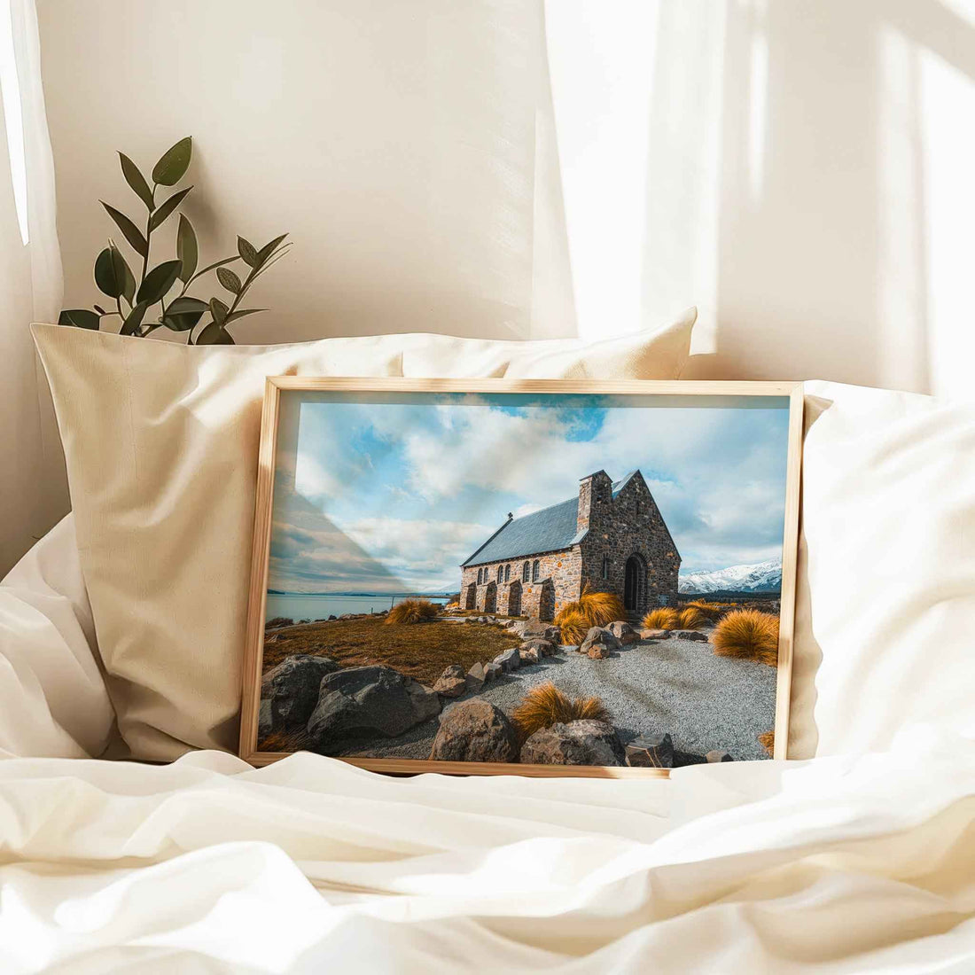 Framed Church of the Good Shepherd Lake Tekapo print in natural wood frame resting against linen pillows on white bed with plant and curtains