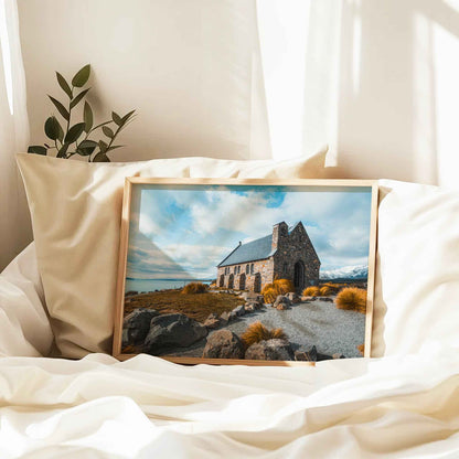Framed Church of the Good Shepherd Lake Tekapo print in natural wood frame resting against linen pillows on white bed with plant and curtains