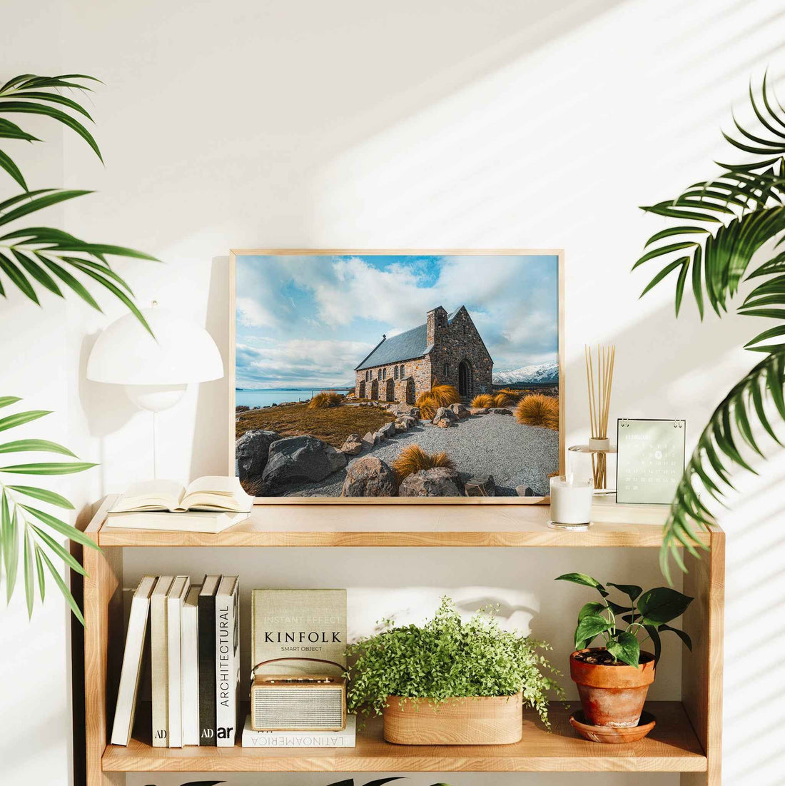 Natural wood framed Church of the Good Shepherd Lake Tekapo print displayed on wooden bookshelf in bright interior styled with plants and books